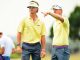 Two LSU golf coaches in yellow shirts stand on a sunny golf course, one pointing as they observe play.
