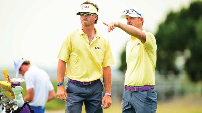 Two LSU golf coaches in yellow shirts stand on a sunny golf course, one pointing as they observe play.