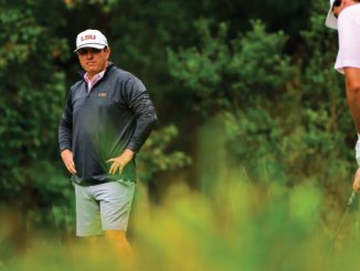Two golfers on a lush course; man wearing an LSU cap and dark jacket stands with hands on hips while another player in a pink shirt crouches with a club furthest right.