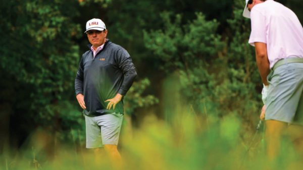 Two golfers on a lush course; man wearing an LSU cap and dark jacket stands with hands on hips while another player in a pink shirt crouches with a club furthest right.