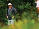 Two golfers on a lush course; man wearing an LSU cap and dark jacket stands with hands on hips while another player in a pink shirt crouches with a club furthest right.