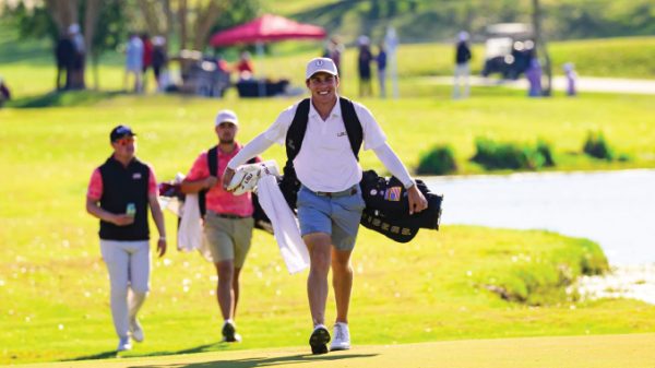 Male golfer walks along a sunny golf course carrying a golf bag over his shoulder, with two caddies following behind.