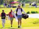 Male golfer walks along a sunny golf course carrying a golf bag over his shoulder, with two caddies following behind.