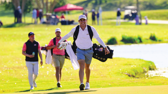 Male golfer walks along a sunny golf course carrying a golf bag over his shoulder, with two caddies following behind.