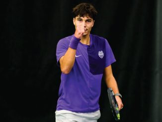 Young male tennis player in a purple shirt, fist raised in celebration, holding a racket on a dark court surface.