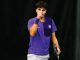 Young male tennis player in a purple shirt, fist raised in celebration, holding a racket on a dark court surface.
