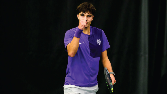 Young male tennis player in a purple shirt, fist raised in celebration, holding a racket on a dark court surface.