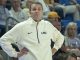 Coach wearing a white LSU jacket with hands on hips, standing on the sideline as spectators watch a game.