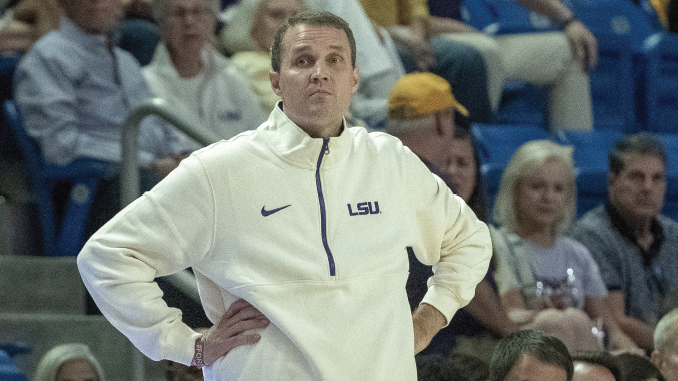 LSUMbkbVMcNeese100826_WillWade_MBacigalupi | TigerRag.com Coach wearing a white LSU jacket with hands on hips, standing on the sideline as spectators watch a game.