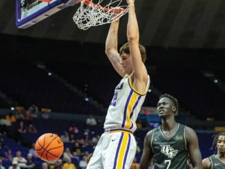 Basketball player in white and yellow jersey dunks the ball at the hoop as a defender watches in a dark arena.