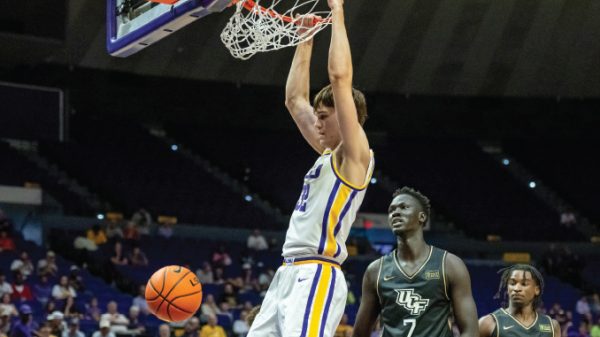Basketball player in white and yellow jersey dunks the ball at the hoop as a defender watches in a dark arena.