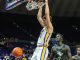 Basketball player in white and yellow jersey dunks the ball at the hoop as a defender watches in a dark arena.