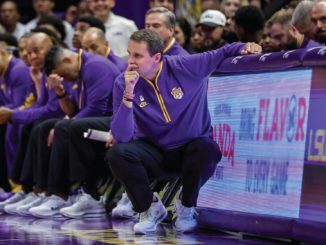 Basketball coach crouched on the sideline, deep in thought, with players in purple uniforms seated behind him.