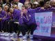 Basketball coach crouched on the sideline, deep in thought, with players in purple uniforms seated behind him.