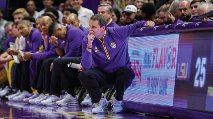 Basketball coach crouched on the sideline, deep in thought, with players in purple uniforms seated behind him.