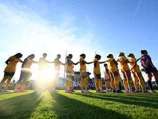 Women soccer players in yellow uniforms form a line on the field, holding hands before a game, bright sun and blue sky.
