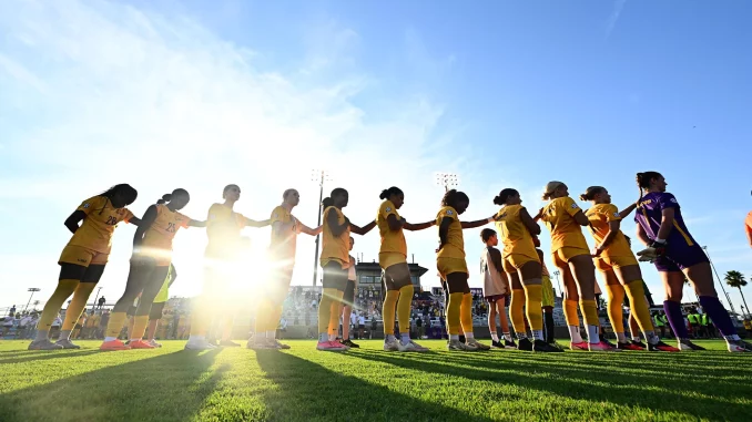 Women soccer players in yellow uniforms form a line on the field, holding hands before a game, bright sun and blue sky.