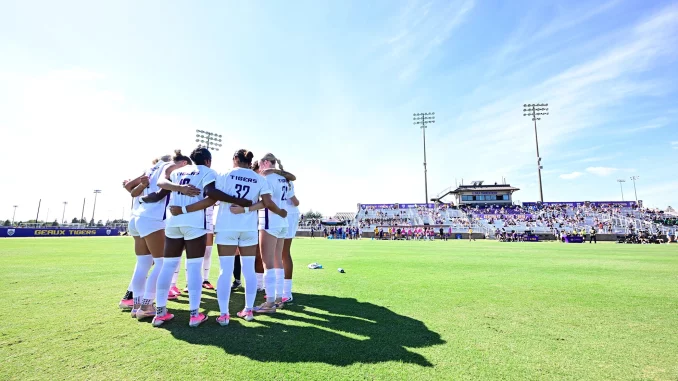 Soccer players in white uniforms huddle on a green field before a game, stands and stadium lights in the background under a blue sky.