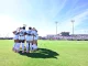 Soccer players in white uniforms huddle on a green field before a game, stands and stadium lights in the background under a blue sky.