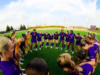 Female soccer players in purple uniforms form a circle huddle on the grassy field before a game.