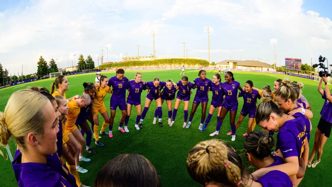 Female soccer players in purple uniforms form a circle huddle on the grassy field before a game.
