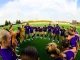 Female soccer players in purple uniforms form a circle huddle on the grassy field before a game.