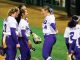 Group of female softball players in purple and white uniforms celebrate on the field, chatting and smiling with a glove visible.