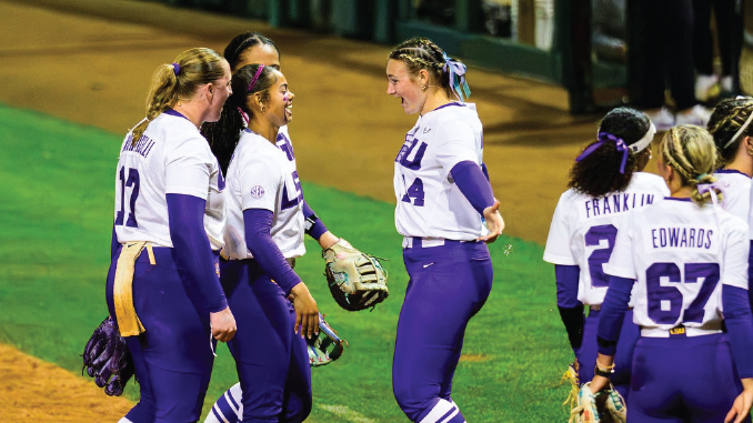 Group of female softball players in purple and white uniforms celebrate on the field, chatting and smiling with a glove visible.