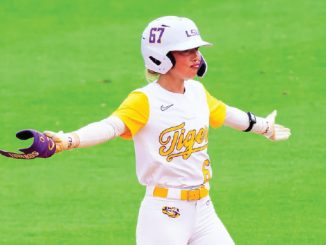 Female softball player in a white and yellow LSU Tigers uniform with helmet on, standing on a green field with arms outstretched as she signals or celebrates a play.