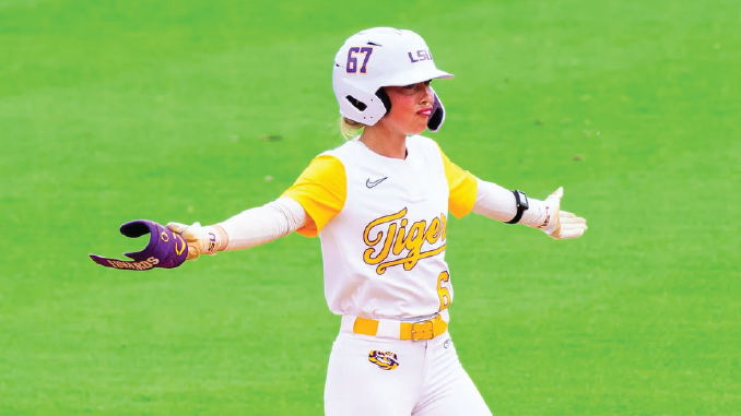 Female softball player in a white and yellow LSU Tigers uniform with helmet on, standing on a green field with arms outstretched as she signals or celebrates a play.
