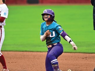 Female softball player in teal jersey with a big 'U' on the chest and purple pants slides into a base, fist raised in celebration as an umpire and opponent look on in the infield.