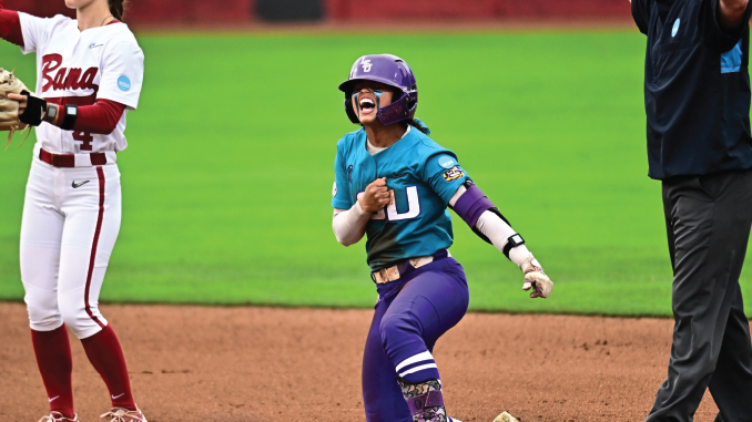 Female softball player in teal jersey with a big 'U' on the chest and purple pants slides into a base, fist raised in celebration as an umpire and opponent look on in the infield.