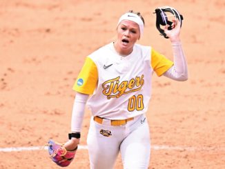 Softball player in a white and gold Tigers uniform (number 00) raises her glove during a game on the orange dirt infield on the mound.
