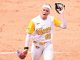 Softball player in a white and gold Tigers uniform (number 00) raises her glove during a game on the orange dirt infield on the mound.