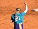Softball player wearing teal jersey with number 21 raises her right hand on a dirt infield, waving goodbye or signaling.