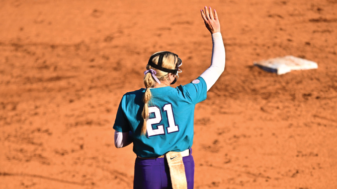 Softball player wearing teal jersey with number 21 raises her right hand on a dirt infield, waving goodbye or signaling.