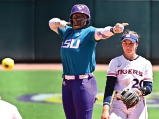 Softball pitcher in teal LSU uniform winds up to throw as a Tigers player in white watches nearby on a sunny field.