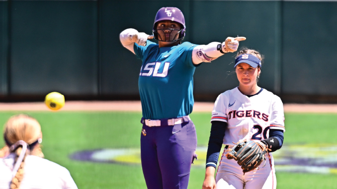Softball pitcher in teal LSU uniform winds up to throw as a Tigers player in white watches nearby on a sunny field.