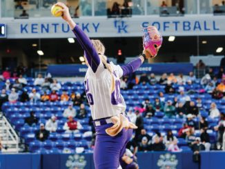 Female softball pitcher in a white and purple uniform winds up to throw on a Kentucky Softball field with spectators in the stands behind her.