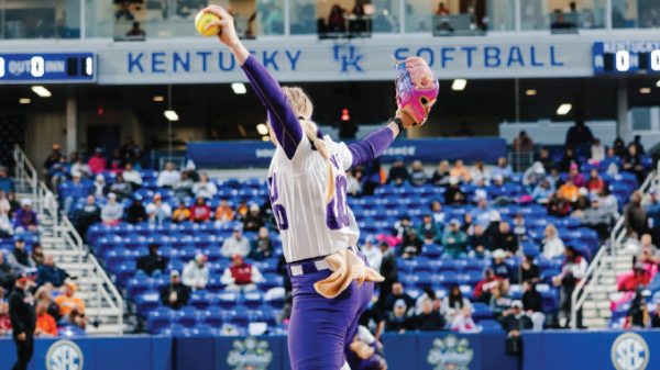 Female softball pitcher in a white and purple uniform winds up to throw on a Kentucky Softball field with spectators in the stands behind her.