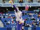 Female softball pitcher in a white and purple uniform winds up to throw on a Kentucky Softball field with spectators in the stands behind her.