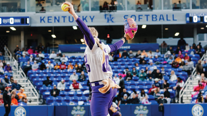 Female softball pitcher in a white and purple uniform winds up to throw on a Kentucky Softball field with spectators in the stands behind her.