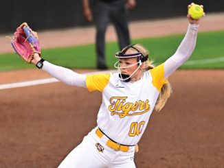 Female softball pitcher in Tigers uniform retracting her throw with arms raised, pink glove on left, holding ball in right hand on the mound.
