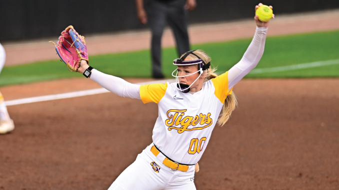 Female softball pitcher in Tigers uniform retracting her throw with arms raised, pink glove on left, holding ball in right hand on the mound.