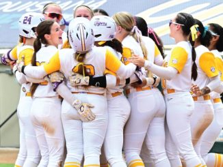 Softball players in yellow and white uniforms hug in a close team huddle on the field, celebrating together.