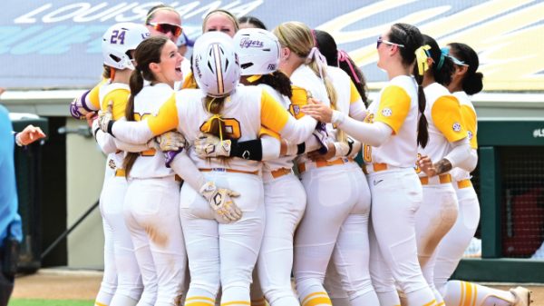 Softball players in yellow and white uniforms hug in a close team huddle on the field, celebrating together.