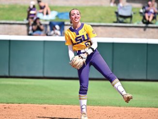 Female softball pitcher in LSU yellow jersey and purple pants delivering a pitch on the infield dirt with fans in the background.
