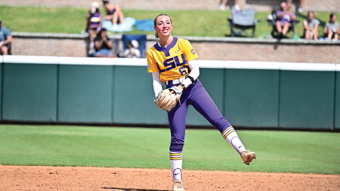 Female softball pitcher in LSU yellow jersey and purple pants delivering a pitch on the infield dirt with fans in the background.