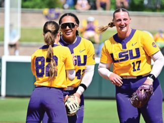 Three LSU softball players in yellow and purple uniforms smile and chat on the field; numbers 84, 14, and 17 visible on their jerseys.