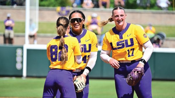 Three LSU softball players in yellow and purple uniforms smile and chat on the field; numbers 84, 14, and 17 visible on their jerseys.